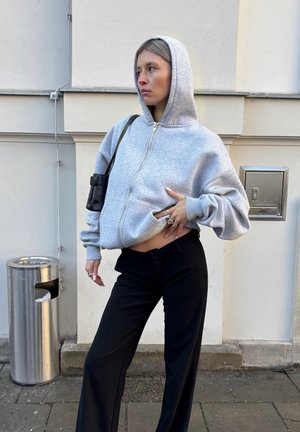 Mujer vestida con una sudadera gris con capucha y cremallera, y pantalones negros, de pie contra una pared blanca con un cubo de basura metálico cerca.