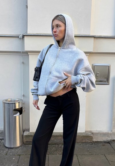 Mujer vestida con una sudadera gris con capucha y cremallera, y pantalones negros, de pie contra una pared blanca con un cubo de basura metálico cerca.