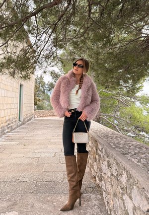 Mujer con chaqueta rosa de peluche, pantalones negros, botas marrones hasta la rodilla y gafas de sol, de pie en un camino de piedra bajo un árbol cerca de una pared de piedra.