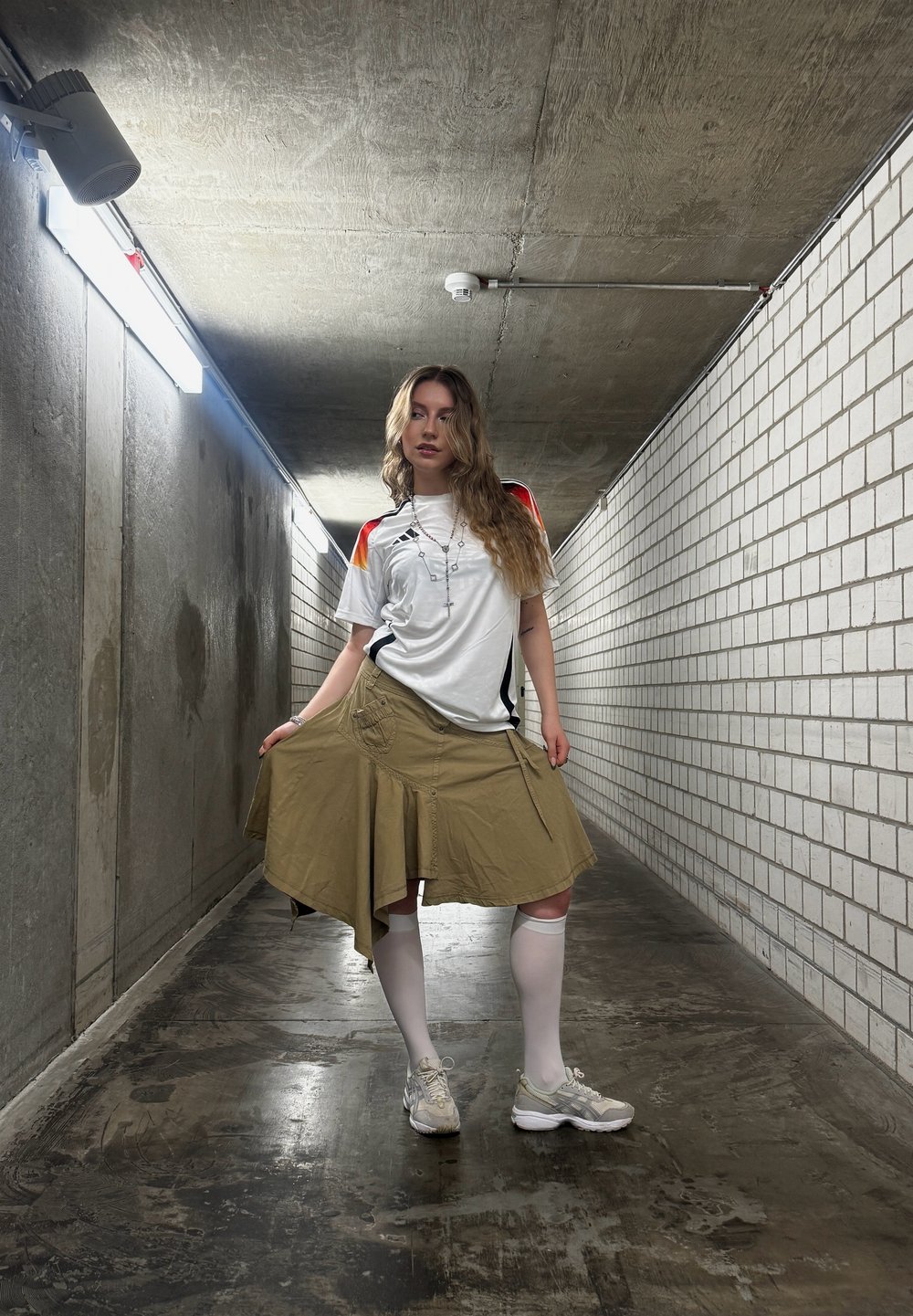 Weisses Sport-T-Shirt mit roten und schwarzen Akzenten, khakifarbenen Wickelrock mit Taschen, weisse knielange Socken und graue Sneakers in einem dämmerigen Flur.