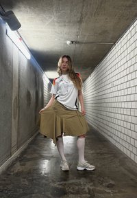 Weisses Sport-T-Shirt mit roten und schwarzen Akzenten, khakifarbenen Wickelrock mit Taschen, weisse knielange Socken und graue Sneakers in einem dämmerigen Flur.
