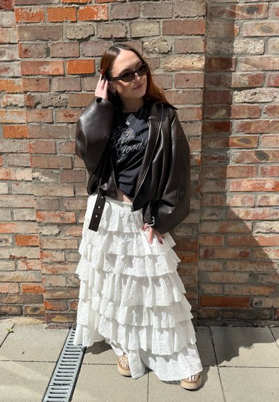 Joven mujer con gafas de sol, chaqueta de cuero negra, top negro, falda blanca de varios volantes y zapatos con estampado de leopardo, parada junto a una pared de ladrillos.