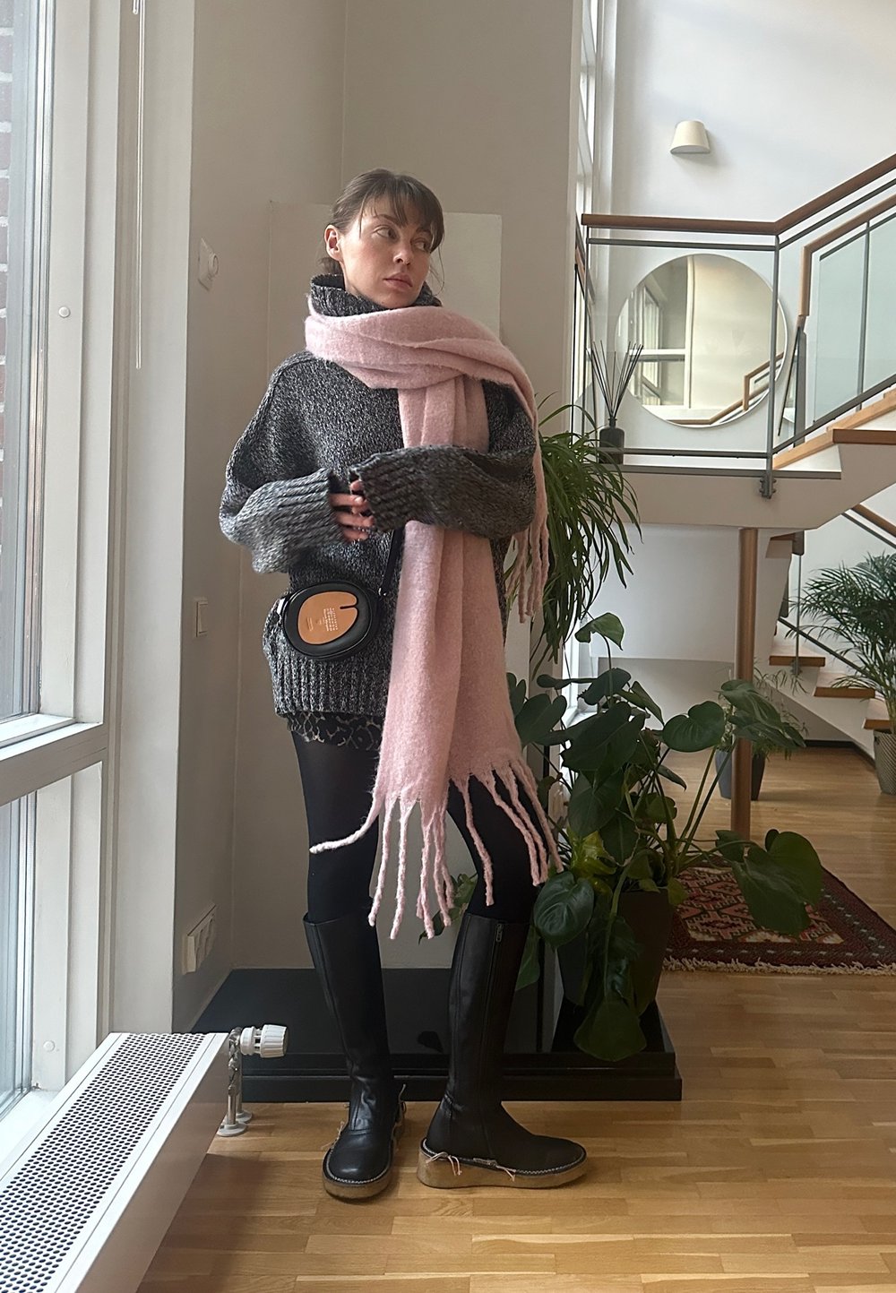Μαύρες μπότες μέχρι το γόνατο, πλεκτή γκρι ζακέτα και μια μακριά ροζ κασκόλ με φούντες. Μαύρη τσάντα χιαστί, εσωτερικά φυτά και ξύλινη σκάλα στο φόντο.
