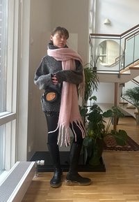 Μαύρες μπότες μέχρι το γόνατο, πλεκτή γκρι ζακέτα και μια μακριά ροζ κασκόλ με φούντες. Μαύρη τσάντα χιαστί, εσωτερικά φυτά και ξύλινη σκάλα στο φόντο.