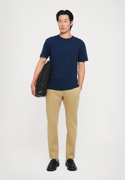 Camiseta de algodón azul marino, pantalones beige ajustados, bolso negro de hombro y mocasines negros, de pie contra un fondo blanco liso.