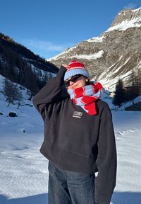 Personne portant un sweat-shirt noir, une écharpe rayée rouge et bleue, un chapeau et des lunettes de soleil, debout dans un paysage montagneux enneigé sous un ciel bleu clair.