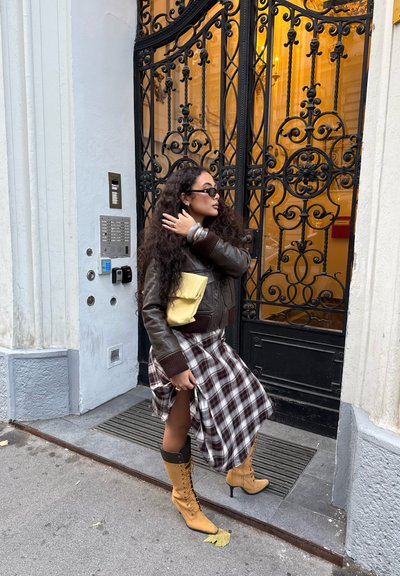 Chaqueta bomber de cuero marrón, falda de cuadros con una abertura, bolso amarillo y botas altas hasta la rodilla de color beige con cordones; puerta ornamentada de hierro forjado de fondo.