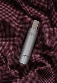 Bouteille de fixateur de maquillage cylindrique argentée sur un tissu pied-de-poule rose. Le texte inclut "MAQUILLAGE VISAGE" et "KIKO MILANO", avec un capuchon métallique lisse.