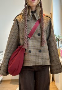 Personne portant une veste croisée marron à carreaux, un pantalon rayé foncé, un bonnet en tricot marron à franges, et un sac bandoulière rouge.