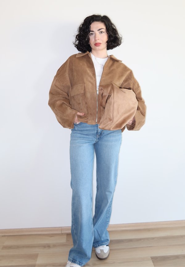 Femme aux cheveux bouclés et foncés portant une veste en daim marron, un jean bleu clair, des baskets blanches, tenant un sac en daim marron assorti.