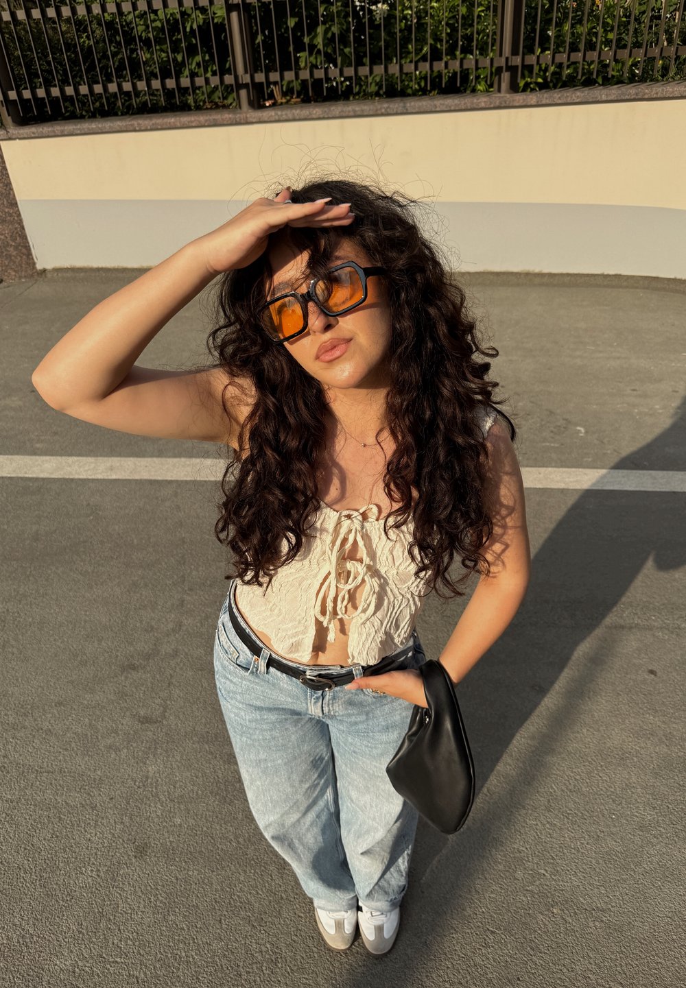 Krem čipkast crop top s vezanjem sprijeda, hlače visokog struka svijetloplave boje, crna torbica, oversize narančaste sunčane naočale i bijeli tenisice.