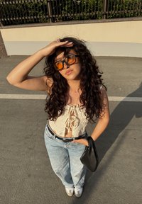 Krem čipkast crop top s vezanjem sprijeda, hlače visokog struka svijetloplave boje, crna torbica, oversize narančaste sunčane naočale i bijeli tenisice.
