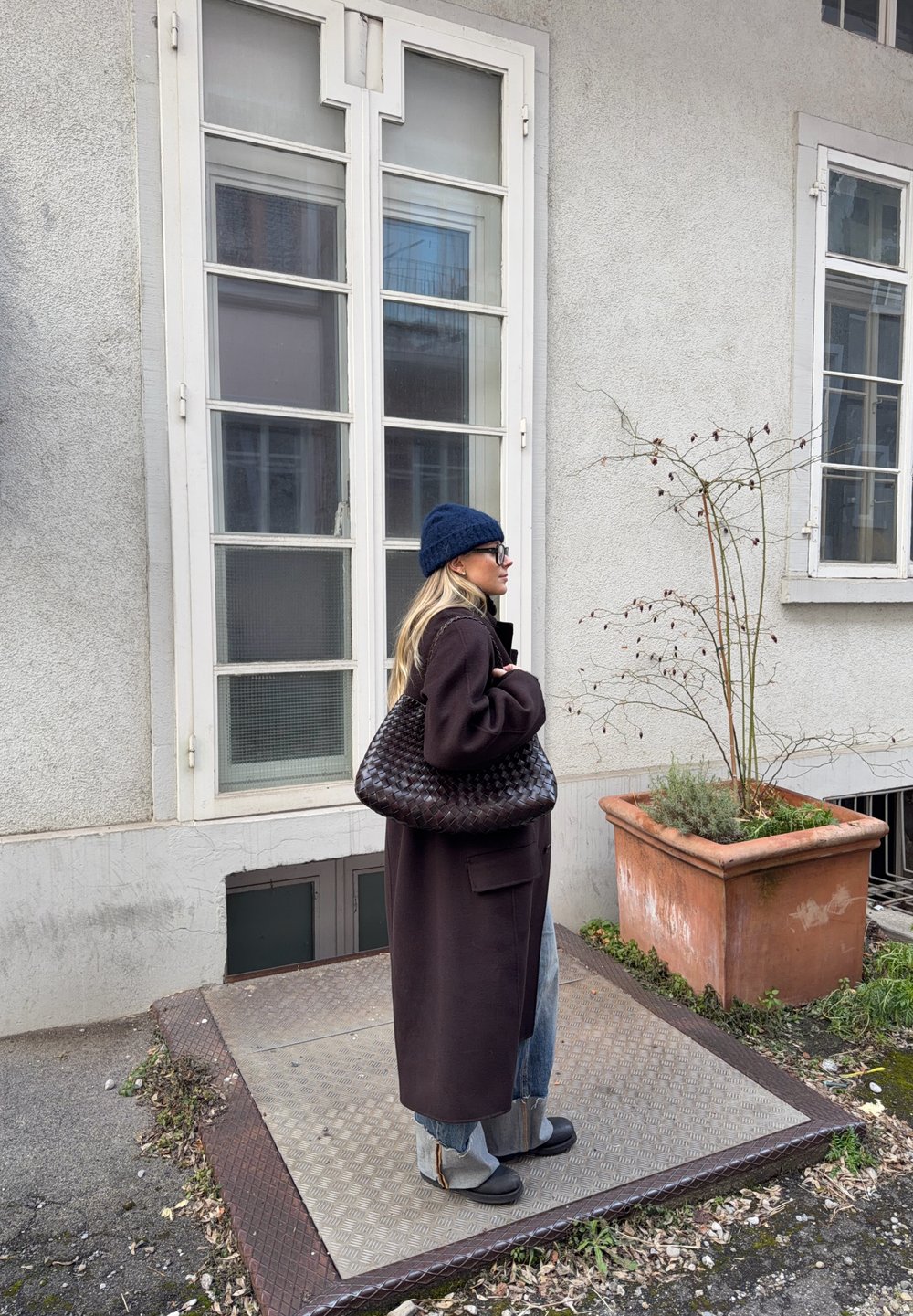 Femme portant un long manteau sombre, un bonnet bleu et des lunettes, debout à côté d'une fenêtre, tenant un grand sac tissé à l'extérieur près d'une plante en pot.