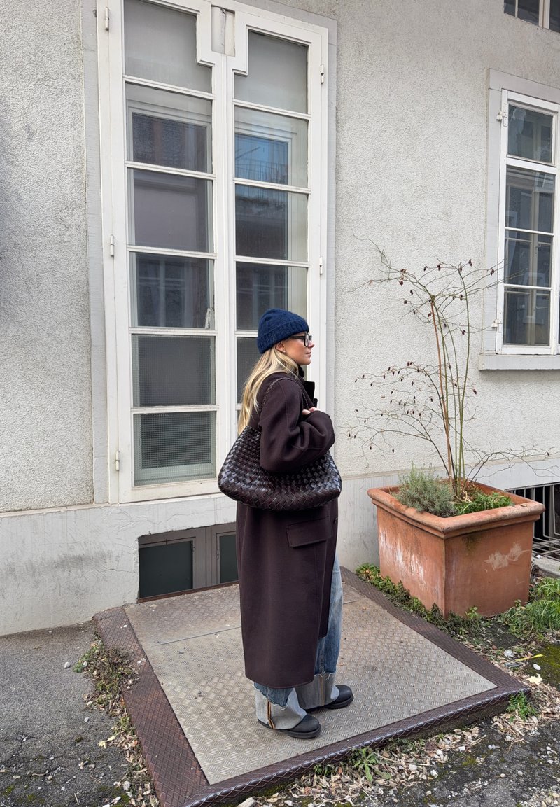 Femme portant un long manteau sombre, un bonnet bleu et des lunettes, debout à côté d'une fenêtre, tenant un grand sac tissé à l'extérieur près d'une plante en pot.