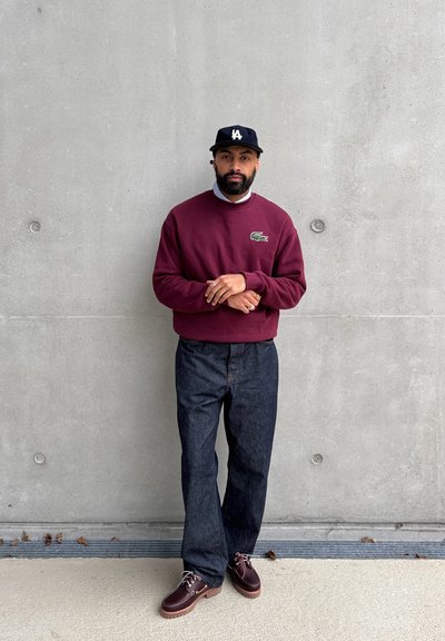 Sudadera burdeos, pantalones de mezclilla oscuros y zapatos burdeos. Gorra negra con logotipo blanco. De pie contra una pared de hormigón gris.