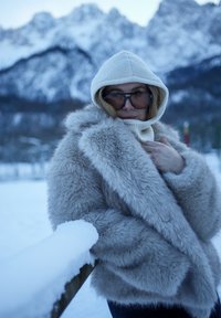 Femme portant de grandes lunettes de soleil, un pull à capuche et un manteau en fourrure duveteuse, debout près d'une clôture en bois couverte de neige avec des montagnes enneigées en arrière-plan.