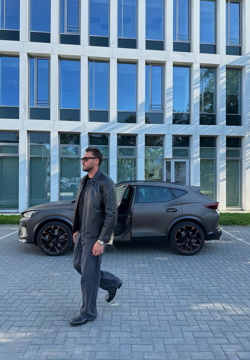Un homme portant une veste en cuir noire et des lunettes de soleil marche devant un SUV gris foncé avec la porte du conducteur ouverte devant un bâtiment moderne en verre.