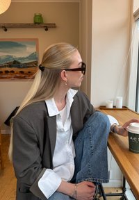 Egy hosszú szőke hajú személy szürke blézerben, fehér ingben és kék farmert visel, zöld kávéscsészét tartva egy faasztalnál, egy ablak mellett.