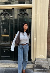 Lys grå cable-knit sweater, højtaljede blå jeans, brunt læderbælte og vævet brun taske, stående foran udsmykkede sorte døre.