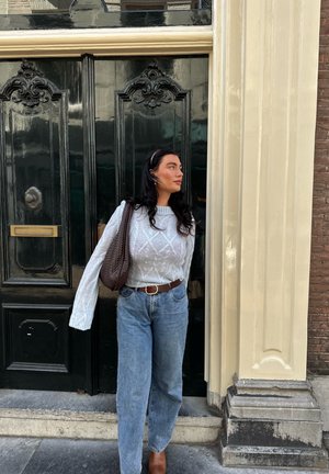 Suéter de punto gris claro, jeans azules de tiro alto, cinturón de cuero marrón y bolso de mimbre marrón, de pie contra puertas negras ornamentadas.