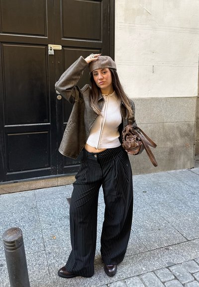 Chaqueta de cuero marrón, top blanco de canalé, pantalones de pierna ancha a rayas blanco y negro, bolso marrón y una gorra marrón claro. De pie al aire libre.