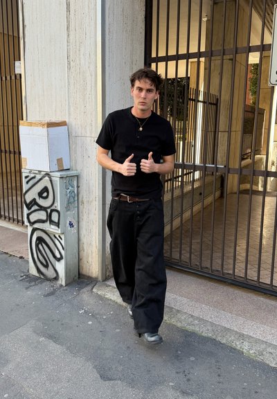 Joven vestido de negro caminando por la acera junto a un edificio vallado, mostrando los pulgares hacia arriba, una caja de cartón sobre una caja de servicios cubierta de grafitis cerca.