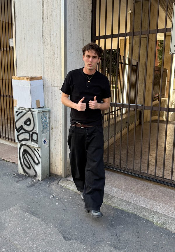 Fiatal férfi fekete ruhában sétál a járdán egy kerítéssel körülvett épület mellett, két hüvelykujját felmutatva, kartondoboz graffitivel borított közműdoboznál a közelben.