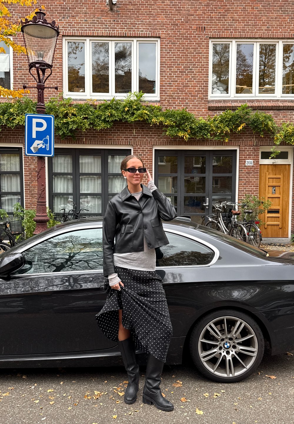 Černá kožená zkrácená bunda, šedé tričko s dlouhým rukávem, sukně s puntíky, černé kozačky po kolena. Zaparkované černé auto s logem BMW v pozadí.