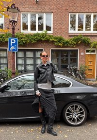Černá kožená zkrácená bunda, šedé tričko s dlouhým rukávem, sukně s puntíky, černé kozačky po kolena. Zaparkované černé auto s logem BMW v pozadí.