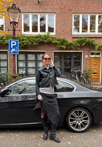Chaqueta de cuero negra corta, camiseta gris de manga larga, falda de lunares, botas negras hasta la rodilla. Coche negro aparcado con el logo de BMW de fondo.