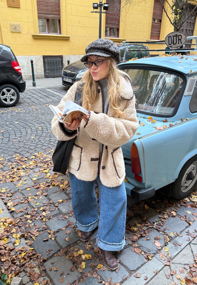 Manteau en polaire beige avec bordure foncée, jeans bleu à jambes larges, bottines marron. Tient un livre ouvert, debout à côté d'une voiture vintage bleue.