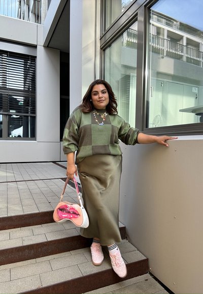 Jersey a cuadros verde y marrón, falda de satén oliva, zapatillas rosa y un bolso brillante con diseño de labios rojos. Entorno urbano al aire libre.