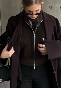 Καφέ σουέτ μπουφάν με φερμουάρ κάτω από ένα σκούρο παλτό, με ορατό κασκόλ. Μαύρη τσάντα με χρυσές λεπτομέρειες, δαχτυλίδια στα δάχτυλα. Λείο υφές.