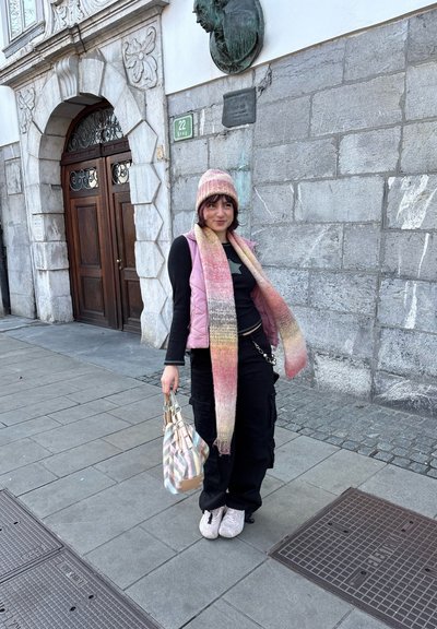 Chaleco acolchado rosa, top negro y pantalones cargo negros y holgados. Bufanda tejida en rosa y naranja. Lleva un bolso de mano multicolor con estampado.