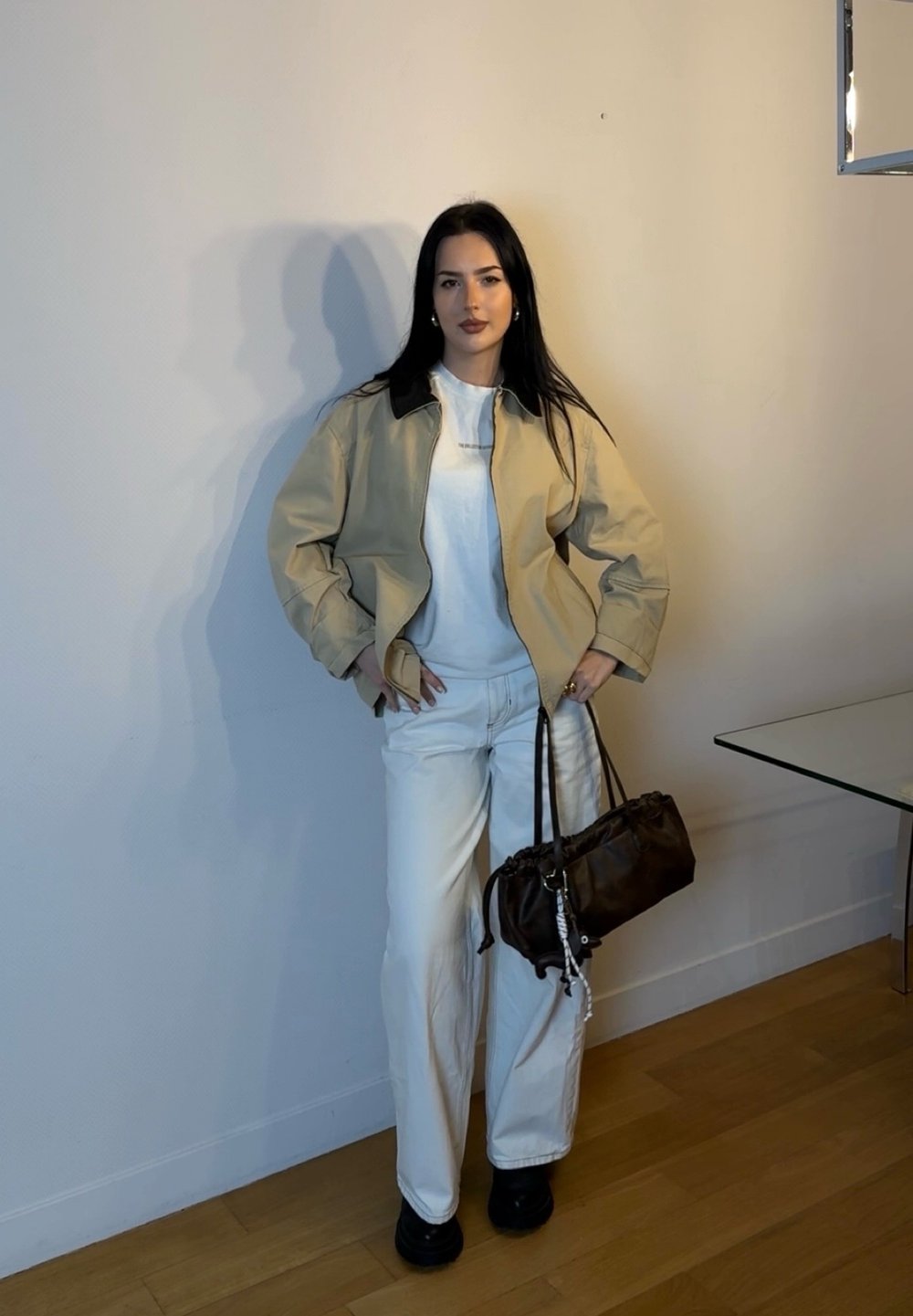 Giacca beige con colletto scuro, camicia bianca e pantaloni bianchi. Il modello tiene una borsa marrone con un design arricciato, scarpe nere, in piedi contro un muro.