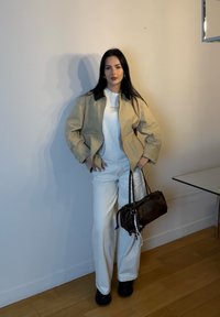 Giacca beige con colletto scuro, camicia bianca e pantaloni bianchi. Il modello tiene una borsa marrone con un design arricciato, scarpe nere, in piedi contro un muro.