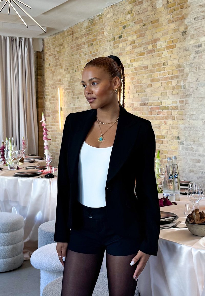 Black blazer over a white bodysuit, paired with black shorts. Braided hair, layered necklaces, and sheer black tights. Elegant dining setting in the background.