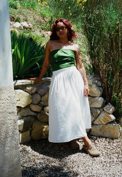 Top sin tirantes verde, falda blanca hasta los tobillos y zuecos marrones. El cabello es ondulado y rojo. De pie junto a una pared de roca con vegetación de fondo.