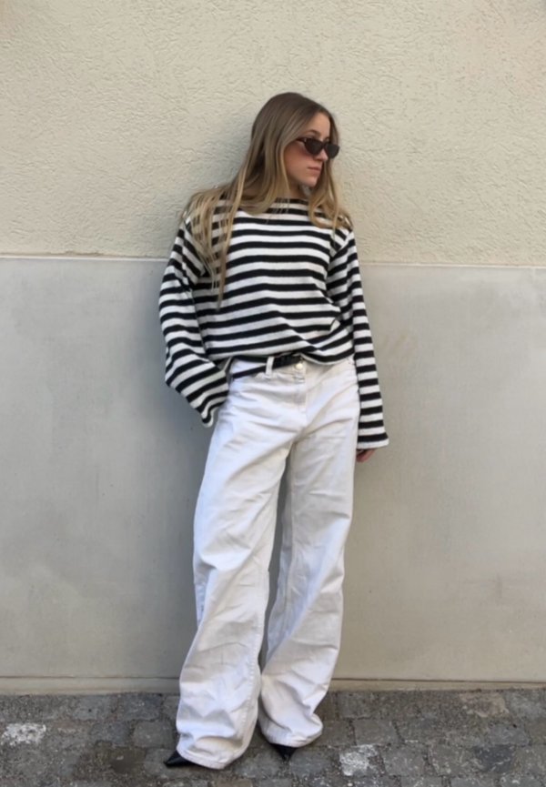 Femme portant un chemisier à manches longues rayé noir et blanc, pantalon blanc à jambes larges, chaussures noires pointues et lunettes de soleil, appuyée contre un mur beige.
