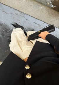 Blazer noir avec boutons en argent, jupe texturée beige, bottes à talons pointues noires et une pochette matelassée noire tenue dans une main.