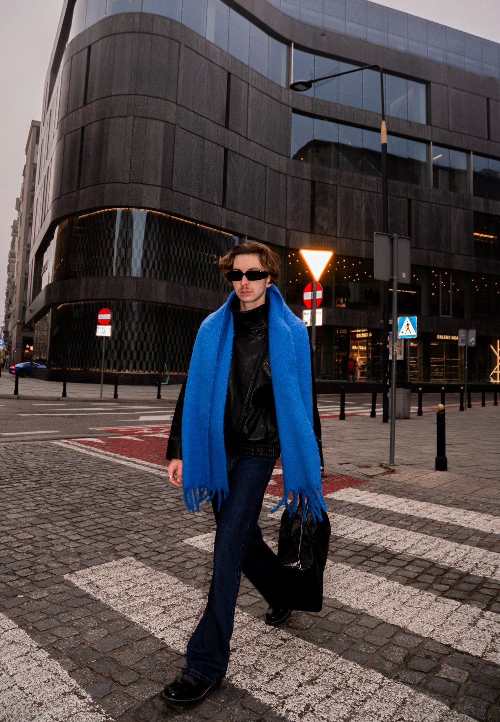 Une personne portant une veste en cuir noire et une écharpe en laine bleue marche sur une rue pavée, portant un sac noir près d'un bâtiment moderne en verre.