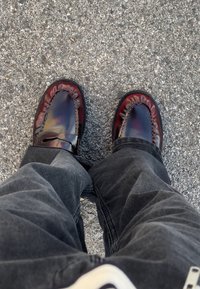 Mocassins de pele castanha com um design de borda franzida, apresentando um detalhe costurado na frente, usados com calças cinza-escuro sobre uma superfície texturizada.