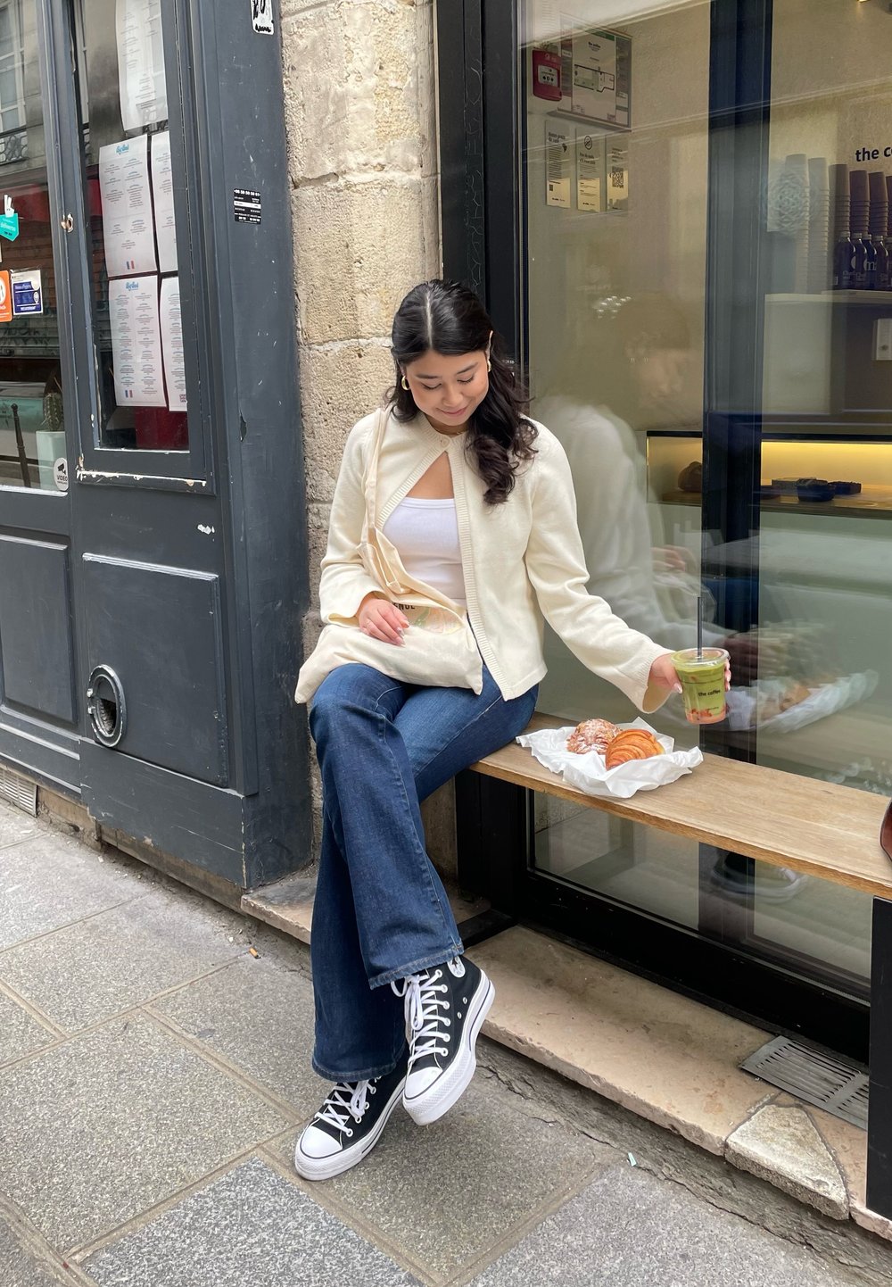 Ανοιχτό μπεζ πλεκτό ζακέτα πάνω από ένα λευκό μπλουζάκι, σκούρα μπλε καμπάνα τζιν και μαύρα και λευκά αθλητικά παπούτσια. Απολαμβάνοντας γλυκά και ένα πράσινο ρόφημα.