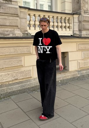 Camiseta negra con el gráfico blanco y rojo "I ❤️ NY", combinada con pantalones holgados negros y zapatos rojos. La persona lleva auriculares.