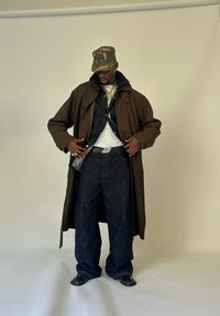 Mann mit Camouflage-Cap, darüber einem mehrlagigen dunklen Trenchcoat über einer Jeansjacke, weißem Hemd, weit geschnittenen Jeans und schwarzen Schuhen vor einfarbigem Hintergrund.