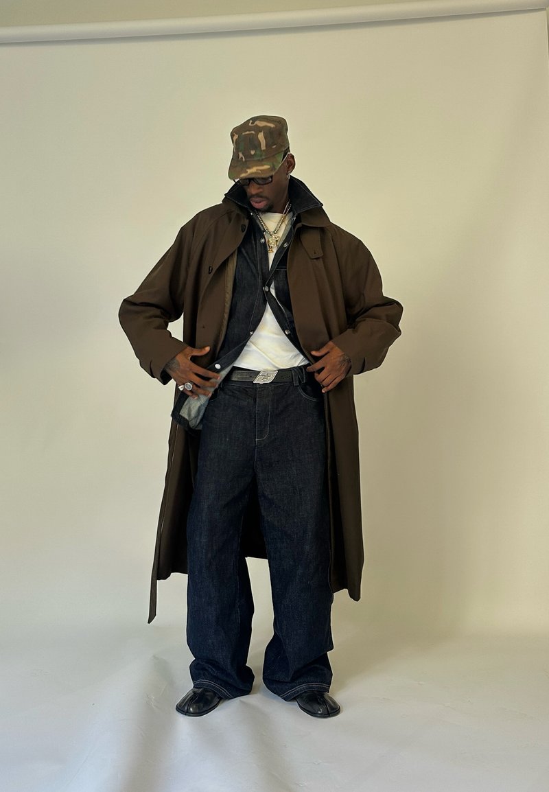 Homem com um boné de camuflagem, um casaco trench escuro em camadas sobre uma jaqueta de denim, camisa branca, calças largas e sapatos pretos em fundo simples.