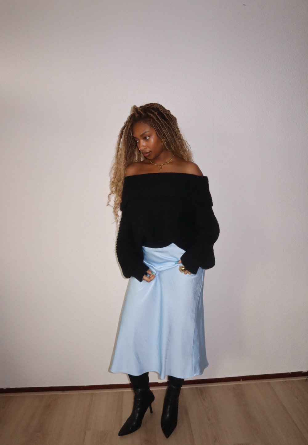 Sweater de malha preto com ombros à mostra, saia de cetim azul claro, botins pretos de salto fino. Texturas suaves, corte descontraído, mangas longas e brilho sutil.