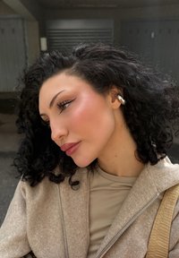 Krøllete mørkt hår, profilbilde. Iført en beige jakke med glidelås og en lys topp. Markant sølvørredobb. Myk sminke med vinget eyeliner.