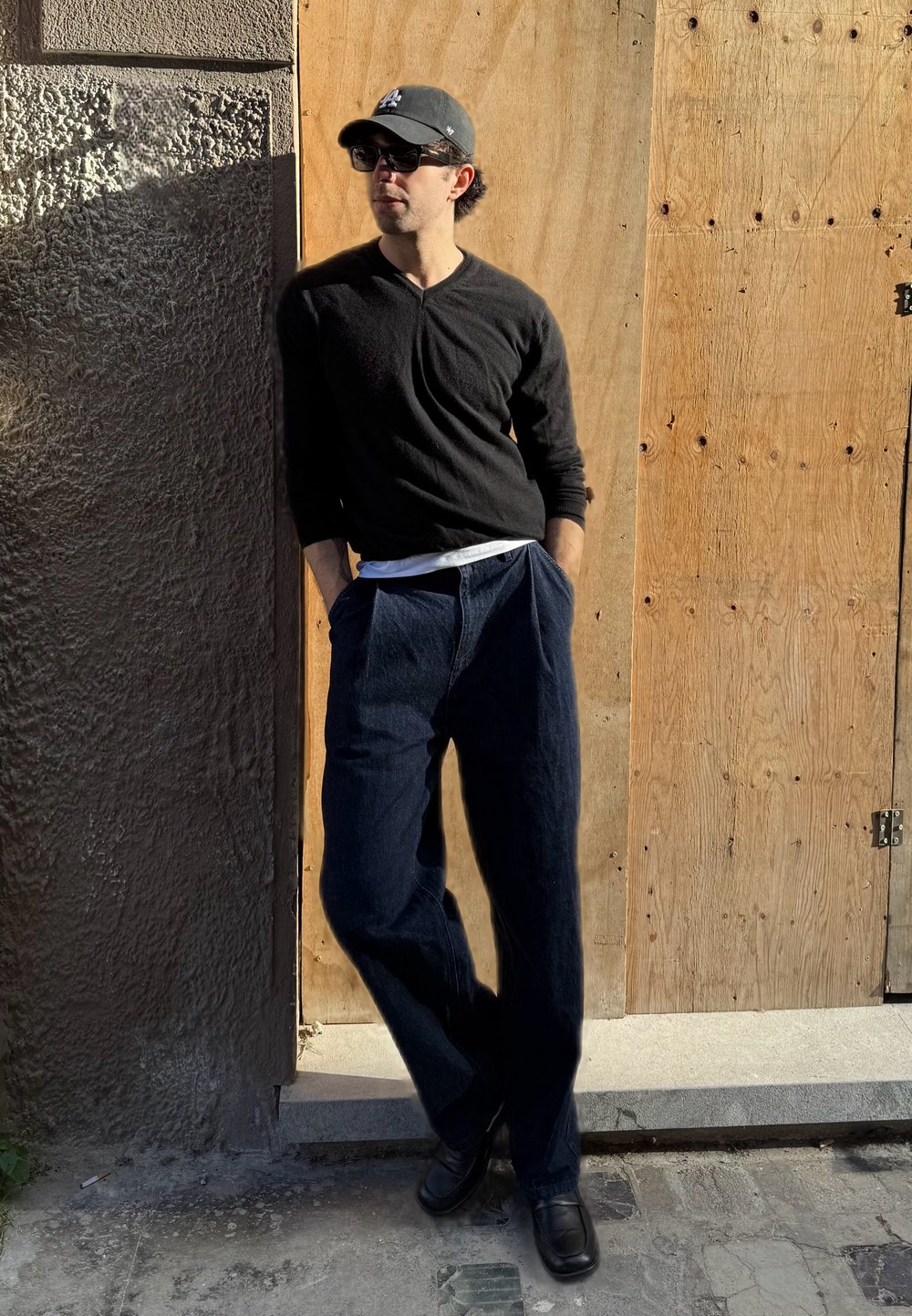 Camisa negra de manga larga y camiseta blanca, pantalones azul oscuro holgados, zapatos negros, de pie contra una pared texturizada y un fondo de madera.
