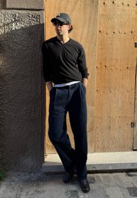 Camisa negra de manga larga y camiseta blanca, pantalones azul oscuro holgados, zapatos negros, de pie contra una pared texturizada y un fondo de madera.